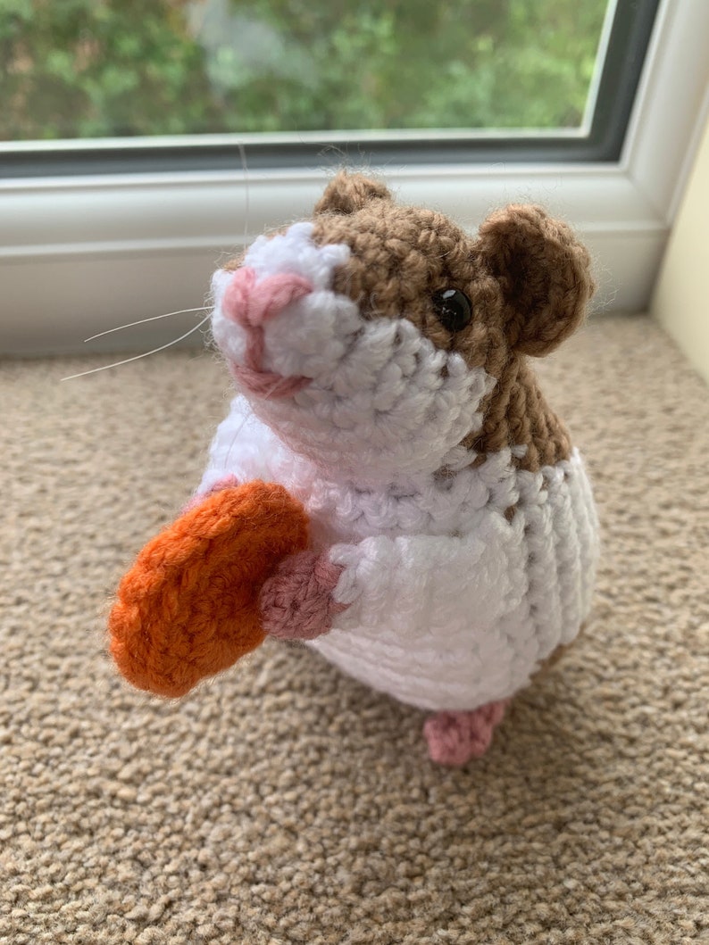 May include: A crocheted hamster toy holding an orange carrot. The hamster is white with brown accents and pink details.