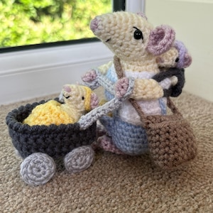 Puede incluir: Una figura de ratón de ganchillo color crema con peto azul y bolso bandolera marrón, empujando un carrito de ganchillo gris oscuro. Dentro del carrito hay un ratón de ganchillo amarillo más pequeño, mientras que otro pequeño se lleva a la espalda.