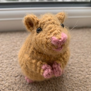 May include: A crocheted hamster toy with brown fur, pink nose, and black eyes. The toy is sitting on a brown carpet.