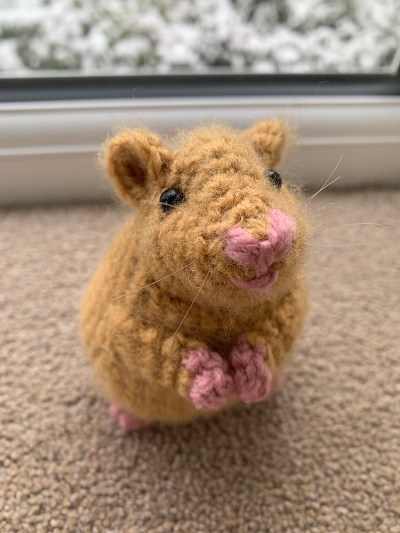 May include: A crocheted hamster toy with brown fur, pink nose, and black eyes. The toy is sitting on a brown carpet.