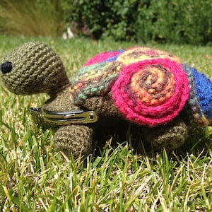 May include: A crocheted turtle toy with a colorful shell. The shell is made of different colored yarn and has a pink, brown, and blue flower design. The turtle is sitting on green grass.