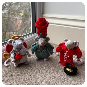Puede incluir: Tres figuritas de ratón de crochet vestidas con diferentes disfraces. Un ratón lleva una corona de rosa roja y una túnica verde, otro lleva un disfraz de corazón rojo y el tercero lleva un halo y alas de ángel.