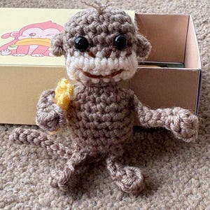 May include: A handmade crochet monkey doll in shades of brown and beige, holding a yellow banana. The monkey has black eyes and a smiling expression. The doll is sitting in front of a box with a monkey graphic.