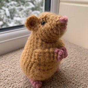 May include: A brown crocheted rat with pink feet and nose. The rat is standing on a brown carpet.