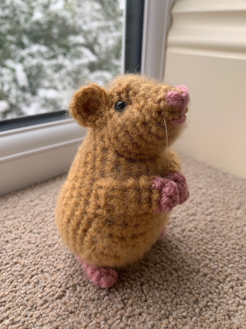 May include: A brown crocheted rat with pink feet and nose. The rat is standing on a brown carpet.