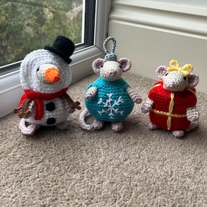 May include: Three crocheted Christmas ornaments: a snowman with a black hat and red scarf, a mouse in a blue sweater with a snowflake, and a mouse in a red coat with a yellow bow. The ornaments are handmade and sit on a carpeted surface.