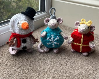 May include: Three crocheted Christmas ornaments: a snowman with a black hat and red scarf, a mouse in a blue sweater with a snowflake, and a mouse in a red coat with a yellow bow. The ornaments are handmade and sit on a carpeted surface.