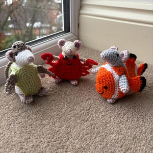 May include: Three handmade crochet toys: a mouse dressed as a possum, a mouse as a crab, and a mouse as a clownfish. The possum mouse is green and brown, the crab mouse is red, and the clownfish mouse is orange, white, and black. All three have pink ears.