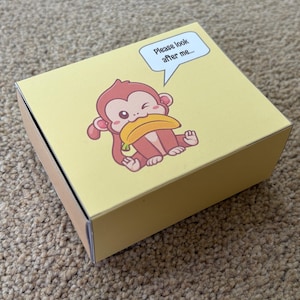May include: A yellow box with a cartoon monkey illustration holding a banana. The monkey is light brown with a winking expression. A speech bubble above the monkey says, "Please look after me..."