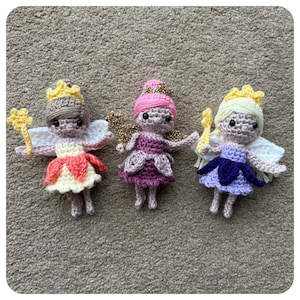 May include: Three handmade crochet fairy dolls. Each doll has a crown, wings, and a wand. The dolls are in shades of pink, purple, yellow, and white. The dolls are on a neutral-coloured surface.