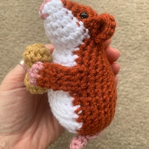 May include: A crocheted hamster toy holding a peanut in its paws. The hamster is brown and white with pink accents.