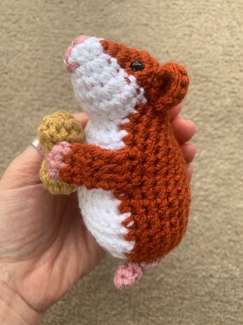 May include: A crocheted hamster toy holding a peanut in its paws. The hamster is brown and white with pink accents.