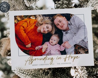 May include: A holiday card featuring a family portrait within a curved frame. The family includes a woman in an orange sweater, a man in a button-down shirt, and a baby. The card reads "Abounding in Hope" and "Merry Christmas 2023".