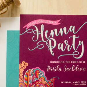 May include: A burgundy invitation with a pink banner that says "Please join us for a Henna Party". The invitation is decorated with a colorful paisley design and the text "Honoring the bride-to-be Prisha Sachdeva" and "Saturday, March 19th 5:00-9:00PM".