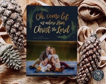 May include: A Christmas card with a blue background and gold lettering that reads "Oh, come let us adore Him Christ the Lord". The card features a family of five with a white dog sitting on a wooden bench. The card is decorated with green leaves and gold glitter.