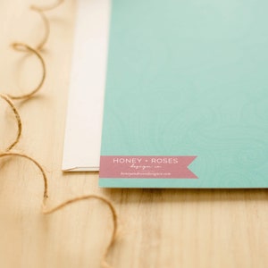 May include: A light blue card with a pink banner that says "Honey + Roses" and a website address. The card is on a wooden surface.