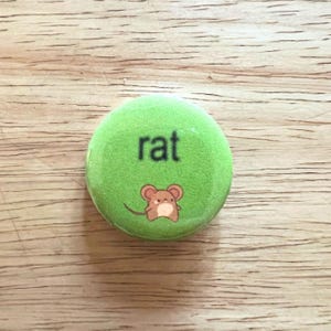 Könnte beinhalten: Ein runder grüner Knopf mit dem Wort "rat" in Schwarz gedruckt. Ein Cartoon-Bild eines braunen Ratten ist auf dem Knopf.