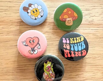 Könnte beinhalten: Fünf bunte runde Knöpfe mit verschiedenen Designs. Die Knöpfe zeigen eine lächelnde Blume, einen Pilz mit einem Smiley, ein Herz mit Beinen, einen Frosch, der Gitarre spielt, und einen schwarzen Knopf mit dem Text "Be Kind To Your Mind" in Regenbogenfarben.