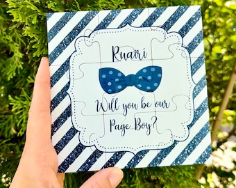 Op de afbeelding: Een wit puzzelstuk met blauwe en witte strepen aan de rand. Het puzzelstuk heeft een blauwe vlinderdas met witte stippen en de tekst "Ruairí Will you be our Page Boy?"