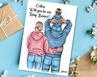 Op de afbeelding: Een puzzelkaart met een illustratie van een bruid en bruidegom in spijkerbroeken en hoodies. De bruidegom draagt een jongetje op zijn schouders. De tekst op de kaart luidt "Cotton, Will you be our Ring Bearer?" en "Love, Auntie Karen & Jacob".