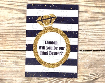 May include: A white and navy blue puzzle card with a gold glitter ring and diamond. The text reads "Landon, Will you be our Ring Bearer?"