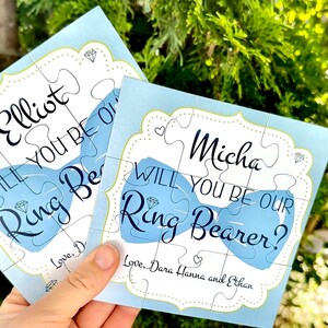 May include: Blue and white puzzle piece card with a bow tie design. The card says "Micha, will you be our ring bearer? Love, Dara Hanna and Ethan".