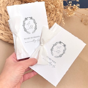 Peut inclure: Deux cartes de mariage blanches avec le texte "TO MY HUSBAND" et "TO MY WIFE" en écriture noire. Chaque carte présente une couronne florale avec les initiales "A M" au centre. Les cartes sont nouées avec des rubans blancs.