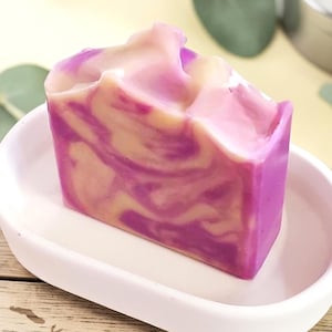 May include: A rectangular bar of soap with a marbled pattern of vibrant purple and creamy white swirls. The soap bar is positioned upright in a smooth, oval-shaped white ceramic soap dish, featuring a textured top edge and a solid base.
