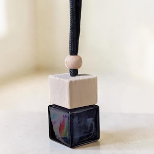 May include: A small square glass bottle with a dark liquid inside, topped with a light-colored wooden cube cap. A single round wooden bead sits above the cap on a black braided cord, allowing the item to be hung for display purposes.