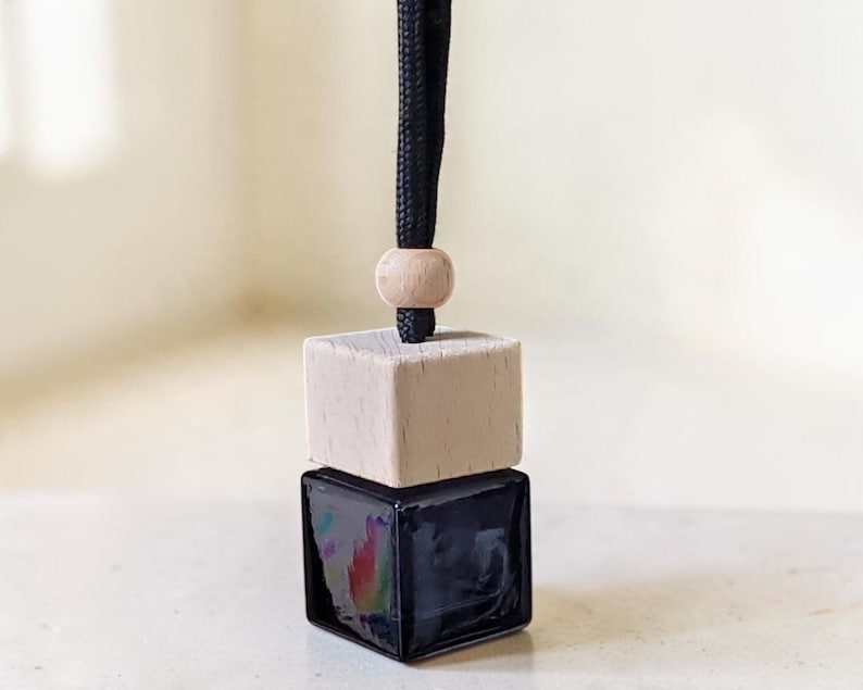 May include: A small square glass bottle with a dark liquid inside, topped with a light-colored wooden cube cap. A single round wooden bead sits above the cap on a black braided cord, allowing the item to be hung for display purposes.