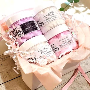 May include: A gift set containing four clear plastic jars of whipped soap with white lids, nestled in light pink tissue paper. The labels read Black Raspberry Vanilla, Coconut Milk & Vanilla, Honeysuckle Strawberry, and Wild Honey Blossom, each 1.5oz.