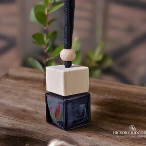 May include: A square glass car air freshener bottle with a dark liquid inside, topped with a light-colored wooden cube cap and a natural wooden bead. A black cord extends from the top for hanging. The text Hickory Ridge Soaps is visible.