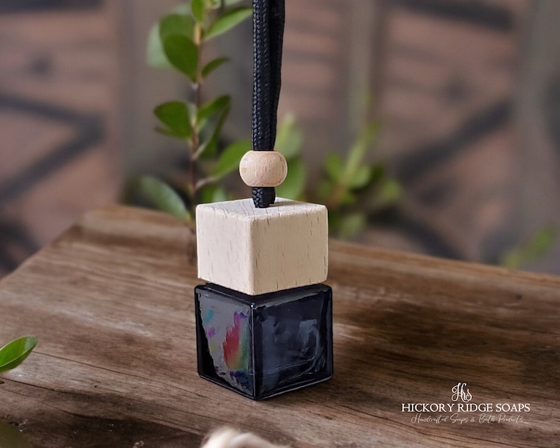 May include: A square glass car air freshener bottle with a dark liquid inside, topped with a light-colored wooden cube cap and a natural wooden bead. A black cord extends from the top for hanging. The text Hickory Ridge Soaps is visible.