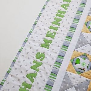 Könnte beinhalten: Eine weiße Babydecke mit grünem und grauem Rand. Die Decke hat ein Muster aus kleinen grauen Herzen auf weißem Grund. Die Worte "Hans Meinhard" sind in Grün auf die Decke gestickt.
