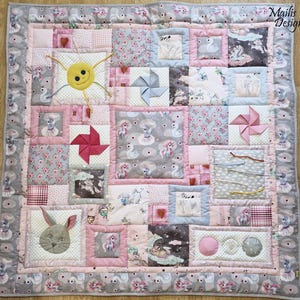May include: A pink and grey patchwork baby play mat with various textures and shapes, including a sun, a bunny, and a windmill. The mat is made of soft fabric and is perfect for babies to explore and play on.