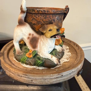 May include: A ceramic dog figurine with brown and white markings, tongue out, rests on a wooden base with a decorative pot. The dog is surrounded by orange flowers and green foliage. The base has a lace doily.