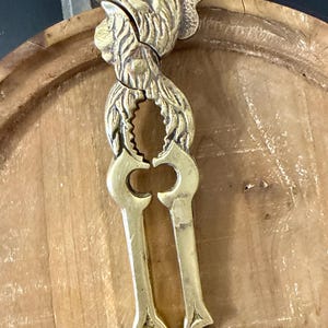 May include: A vintage brass nutcracker with a detailed rooster head design. The nutcracker features a lever mechanism and is made of brass. The rooster's head is intricately detailed, adding a decorative element to the functional tool.