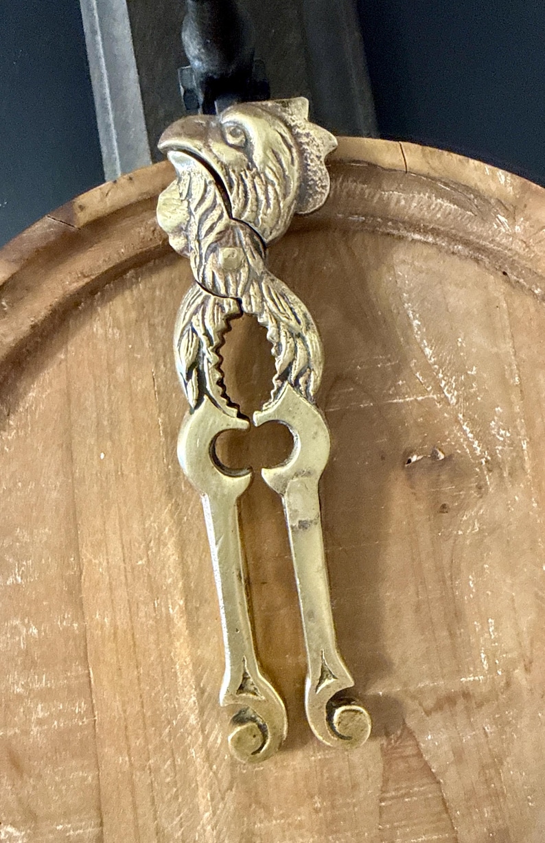 May include: A vintage brass nutcracker with a detailed rooster head design. The nutcracker features a lever mechanism and is made of brass. The rooster's head is intricately detailed, adding a decorative element to the functional tool.