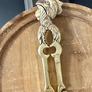 May include: A brass nutcracker shaped like a rooster. The nutcracker is gold-colored and features a detailed design of a rooster's head and body. The handles are long and have decorative cutouts. The nutcracker is resting on a wooden surface.
