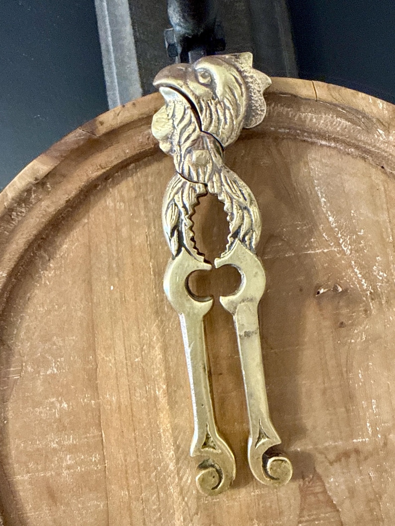 May include: A brass nutcracker shaped like a rooster. The nutcracker is gold-colored and features a detailed design of a rooster's head and body. The handles are long and have decorative cutouts. The nutcracker is resting on a wooden surface.