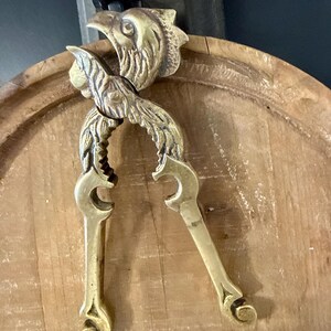 May include: A vintage brass nutcracker with a detailed eagle head design. The nutcracker is gold-colored and features a lever-style mechanism with ornate, curved handles. The eagle's head is at the top, and the handles are curved.