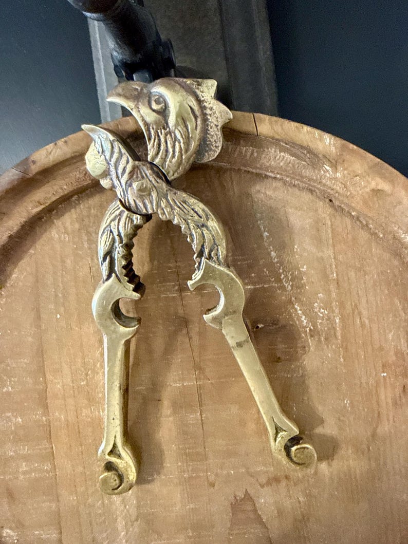 May include: A vintage brass nutcracker with a detailed eagle head design. The nutcracker is gold-colored and features a lever-style mechanism with ornate, curved handles. The eagle's head is at the top, and the handles are curved.