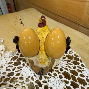 May include: A ceramic salt and pepper shaker set featuring a yellow hen with a red comb and beak, flanked by two smooth, glossy, golden-yellow egg-shaped shakers. The hen has textured feathers and dark brown feet, with the shakers held at its sides.