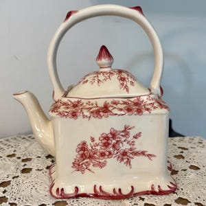 May include: Cream-colored ceramic teapot with a square body and a curved handle. The teapot is decorated with a red floral pattern and trim. The lid has a red finial. The teapot is sitting on a white lace tablecloth.