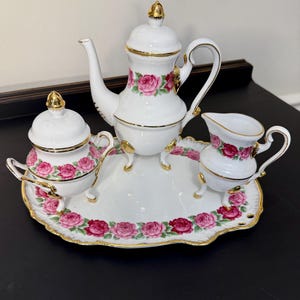 May include: A white porcelain tea set with pink rose floral pattern and gold trim. The set includes a teapot, sugar bowl, creamer, and a serving tray. The teapot has a lid with a gold finial and sits on three gold feet.