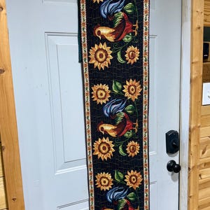 May include: A long, rectangular fabric table runner featuring a black background with a repeating pattern of colorful roosters and yellow sunflowers. The runner has a decorative border with small floral motifs and is hanging on a white hanger.