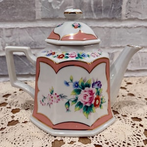 May include: A white porcelain teapot with a pink and gold trim, featuring floral designs. The teapot has a lid and a handle. The floral design includes pink, red, blue, and purple flowers. The teapot is sitting on a lace doily.