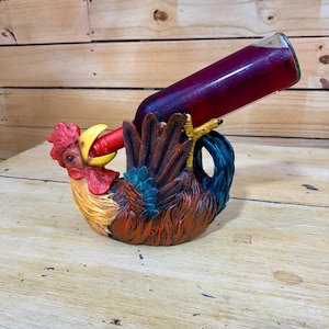 May include: A rooster-shaped wine bottle holder featuring a painted finish with red, yellow, and blue feathers. The sculpture depicts a rooster with an open beak supporting a glass bottle filled with a dark red liquid at an angle.