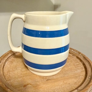 May include: Cream-colored ceramic pitcher with horizontal blue stripes. The pitcher has a curved handle and a spout for pouring. It is sitting on a round, wooden tray.