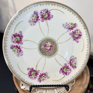May include: A white porcelain plate with a floral design. The plate features clusters of purple and green flowers arranged around the rim and center. A gold-colored trim and accents complete the design. The plate is displayed on a black stand.